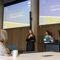 Two students speaking to a group, with the words "NICE summer school" on projectors behind them.