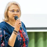 A person wearing a traditional Ukranian blouse speaking with a microphone. A projector screen is visible behind them.