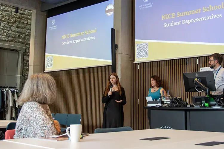 Two students speaking to a group, with the words "NICE summer school" on projectors behind them.