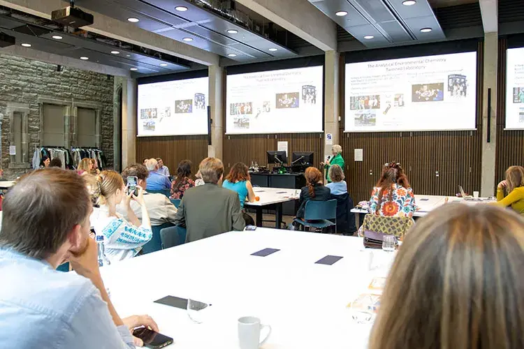 A group of people sitting at tables in a classroom looking up at projector screens.