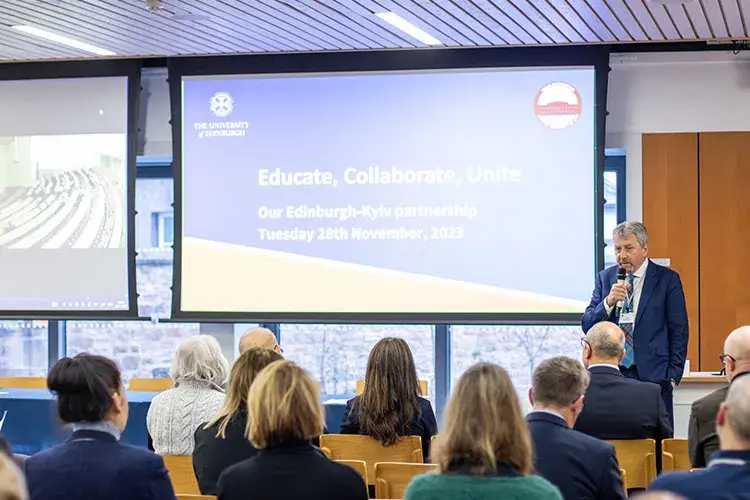 A person in a suit speaking in front of a small group of people. A projector reads "Educate, Collaborate, Unite".
