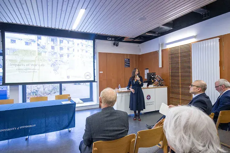 A person in a classroom standing in front of a projector screen speaking to a group.