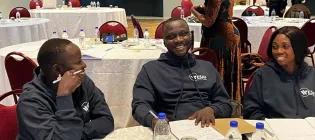 Three Mastercard Foundation Scholars sitting and laughing at an event. Several large round tables are in the background.